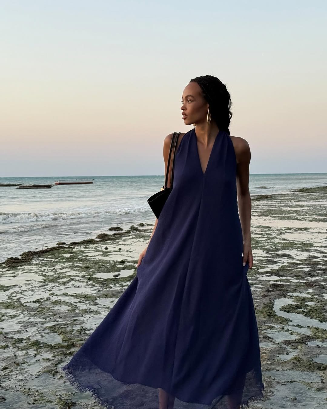 Vestidos midi para o verão são perfeitos para ocasiões à beira-mar. Nesta imagem, uma mulher usa um vestido midi azul marinho, com decote em V, que flui suavemente enquanto ela caminha na areia. A combinação com acessórios simples, como uma bolsa preta e