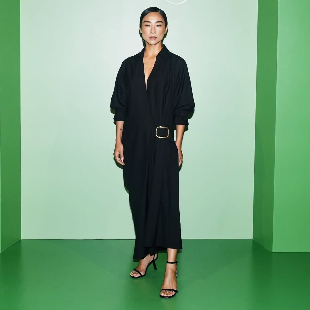 Greta Lee exibe um elegante vestido preto de corte longo, com um decote profundo e mangas bufantes, que traz um toque contemporâneo à peça. O cinto largo em destaque acentua a silhueta, enquanto as sandálias de tiras finas complementam o visual. O fundo v