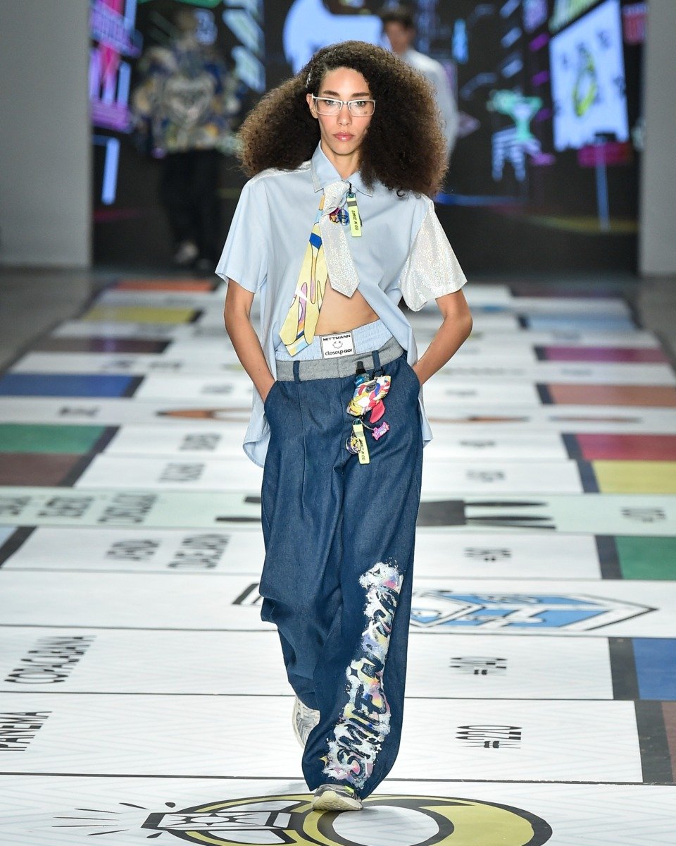No desfile, uma modelo exibe um conjunto que combina uma camisa azul clara e calças jeans amplas, adornadas com detalhes coloridos. O look é complementado por um chaveiro estiloso preso à  calça, adicionando um toque de personalidade.