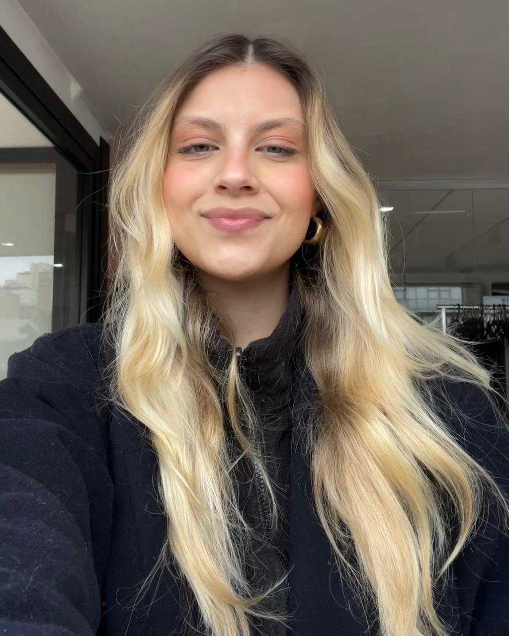 Mulher sorrindo com maquiagem em blush terracota e cabelo loiro comprido, usando casaco preto.