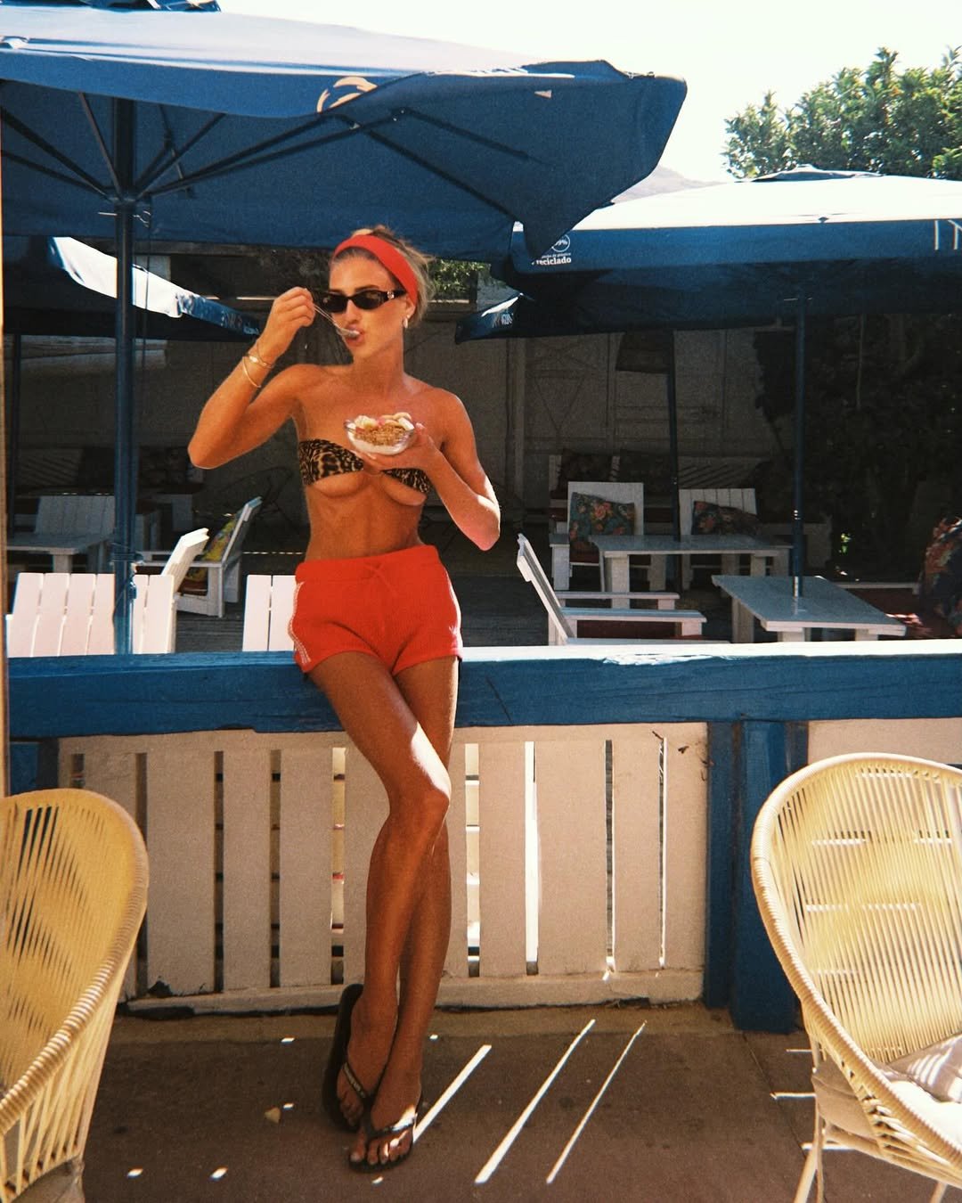 Mulher em resort à beira-mar, usando top estampado e shorts vermelhos. O estilo evoca moda surfista.