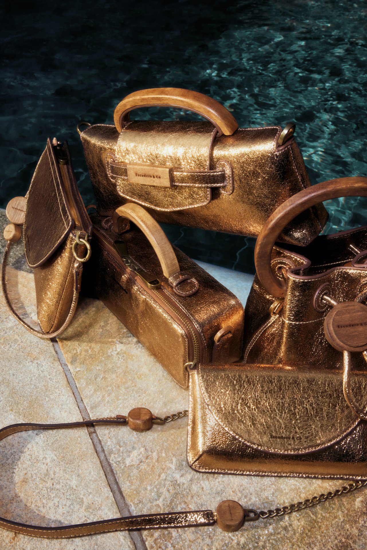 Bolsas douradas com detalhes em madeira dispostas sobre uma borda de piscina. Cápsula Âmbar