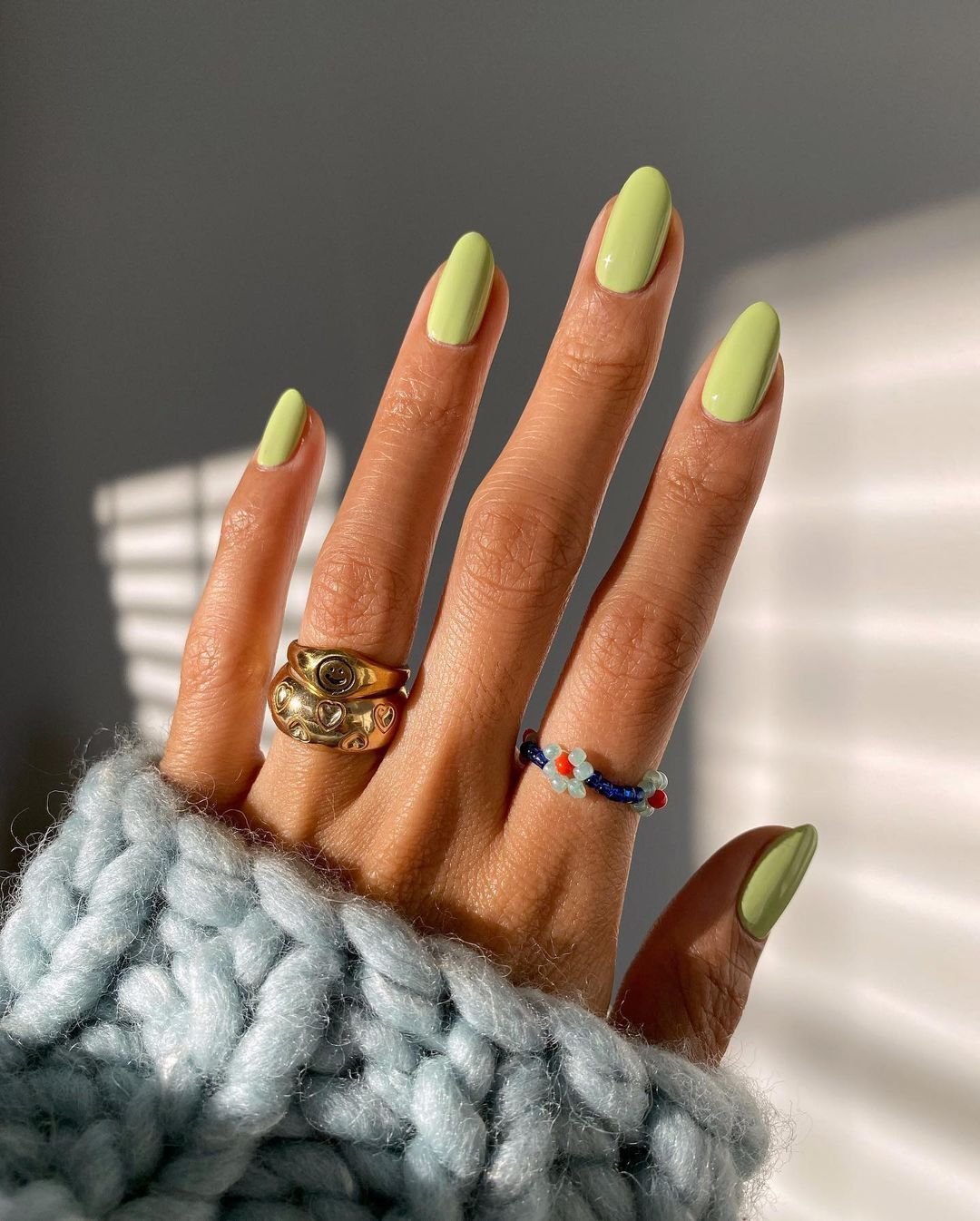 Unhas com esmalte verde claro, anéis dourados e de miçangas, blusa de tricô azul, representando esmalte primavera verão 2026.
