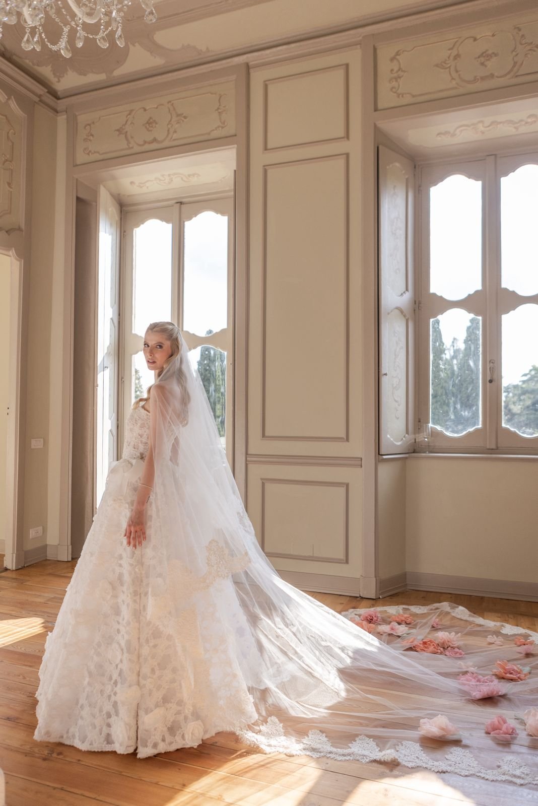 Noiva em vestido elegante com cauda longa decorada, em ambiente clássico iluminado.