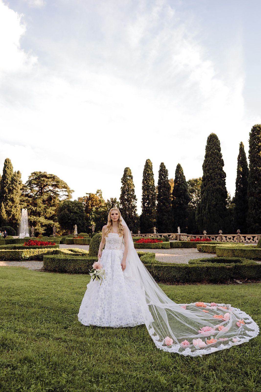 Noiva em vestido de casamento luxuoso, com véu floral, desfilando em jardim elegante.