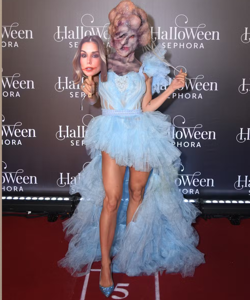 A imagem apresenta uma fantasia de Halloween inovadora, combinando um vestido de tule azul claro com detalhes dramáticos. A peça é curta na frente e longa atrás, conferindo um toque elegante. A maquiagem e a máscara de rosto adicionam um elemento surreal,
