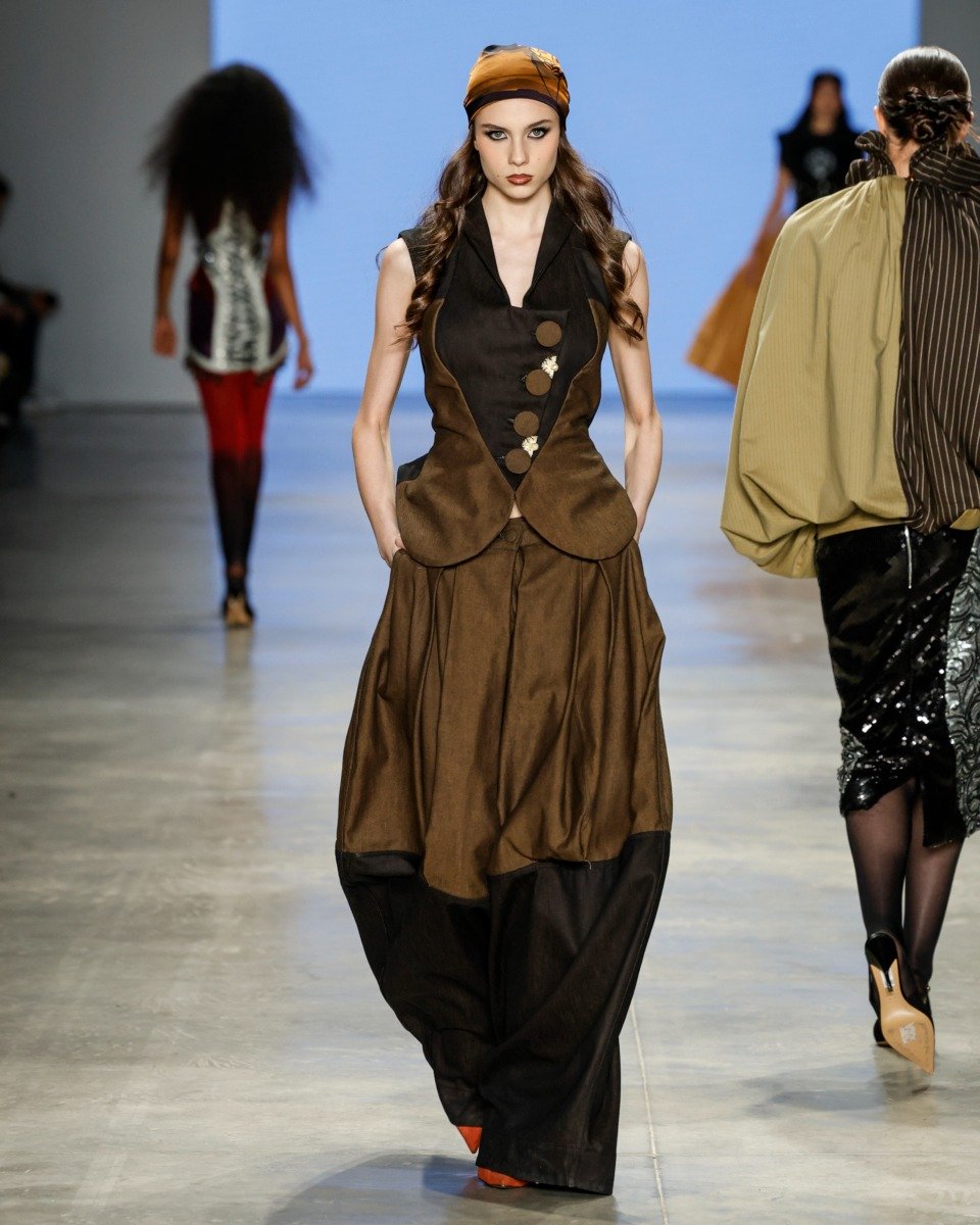 Na SPFW N60, uma modelo desfila com um look sofisticado, destacando um vestido em tons terrosos, com um colete de botões que valoriza a silhueta. O cabelo está preso com um lenço, complementando o estilo contemporâneo. Ao fundo, outras modelos exibem peça
