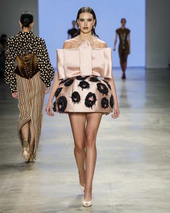 No SPFW N60, uma modelo exibe um vestido ousado com uma saia volumosa em tom rosa claro, adornada com flores escuras. A parte superior apresenta um design estruturado, destacando ombros.