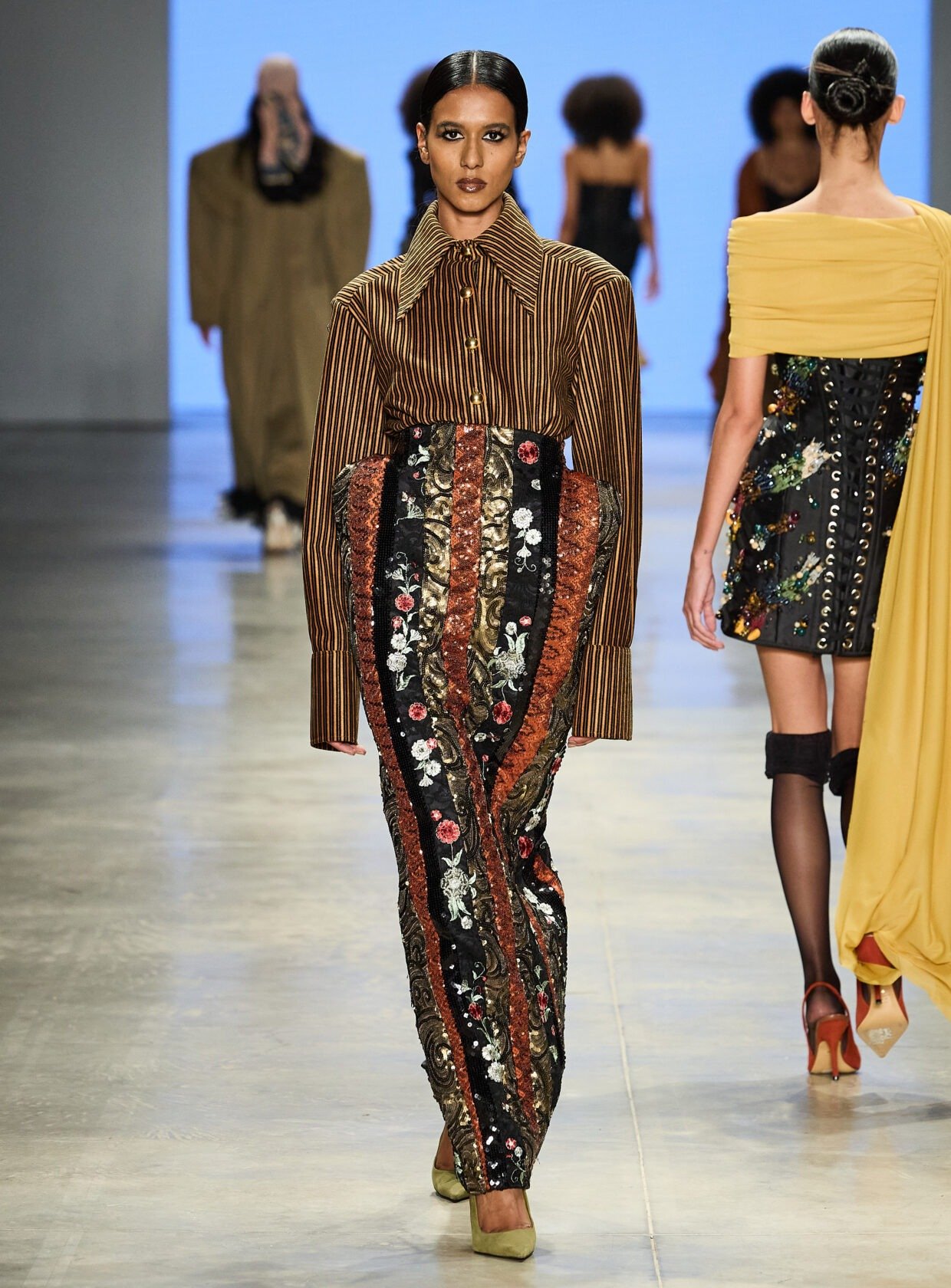 No SPFW N60, uma modelo destaca-se na passarela com um look ousado. A blusa listrada de gola ampla combina com uma saia longa ricamente bordada, mesclando tons de preto, vermelho e detalhes florais.