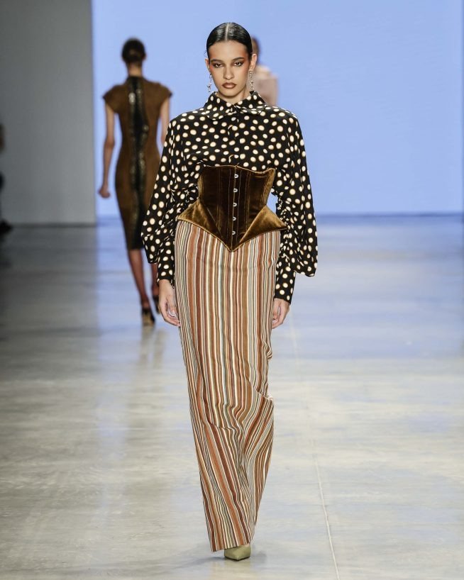 Modelo desfila na SPFW N60, usando uma blusa de bolinhas preta e branca, com mangas bufantes, combinada a uma saia listrada em tons terrosos. O destaque fica para o corset em veludo marrom, que acentua a silhueta.
