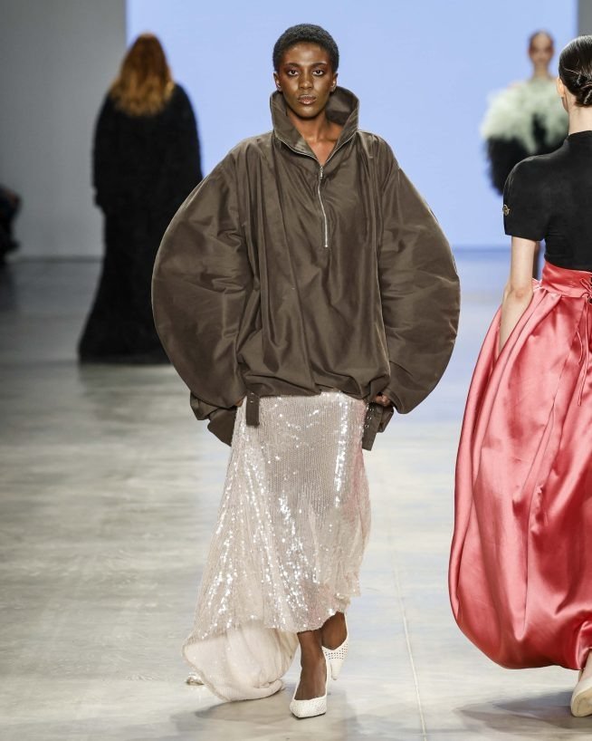 No SPFW N60, uma modelo desfila com um look impactante que combina uma blusa oversized em tom marrom e uma saia longa com paetês prateados. O contraste entre a silhueta volumosa e a fluidez da saia cria um efeito visual marcante.