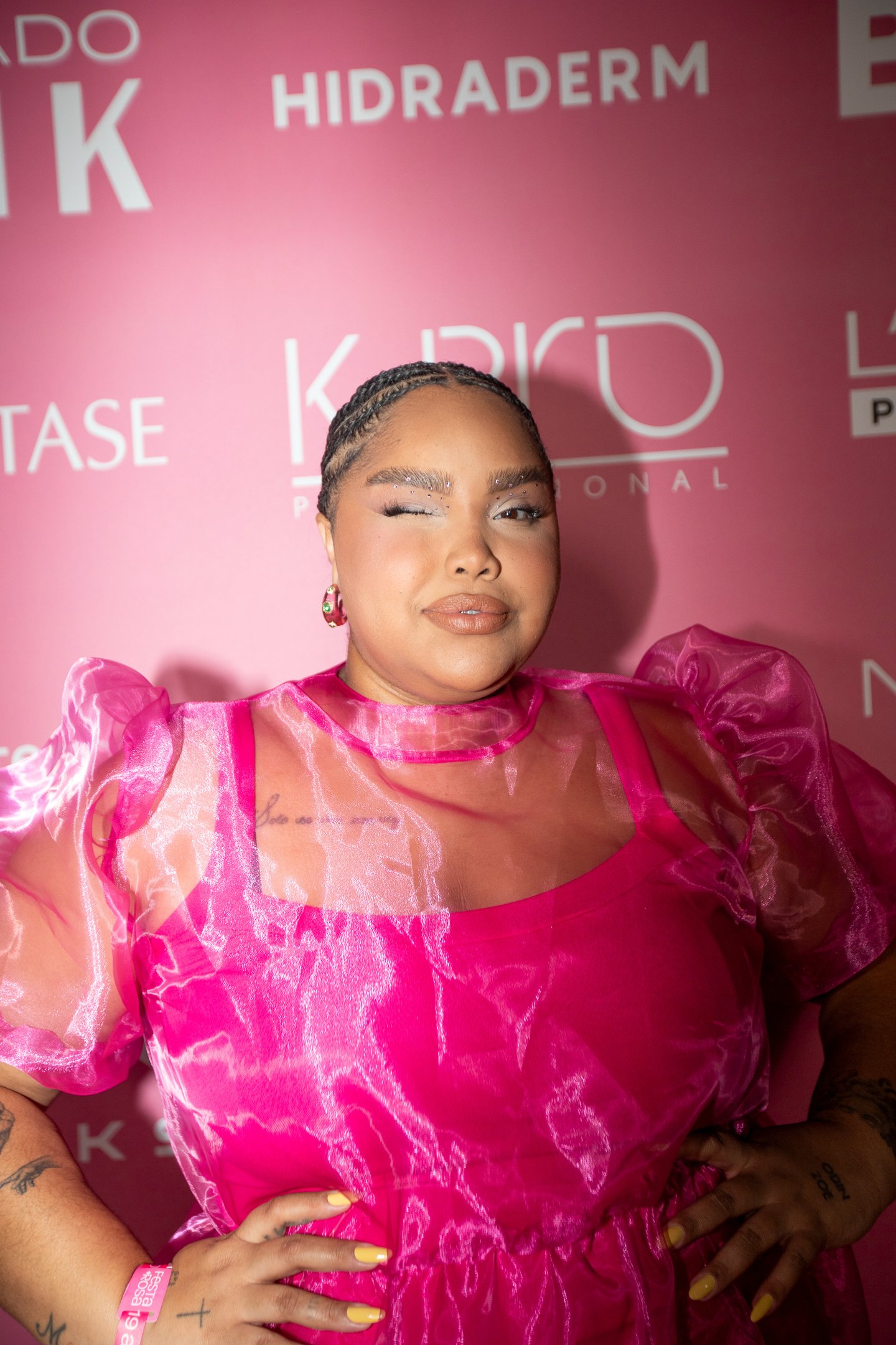 Na Festa do Rosa, uma mulher exibe um look vibrante com um vestido pink de mangas bufantes, que combina transparência e brilho. Seu cabelo está trançado, e ela usa maquiagem com destaque para os olhos e lábios. O cenário rosa ao fundo reforça a temática d