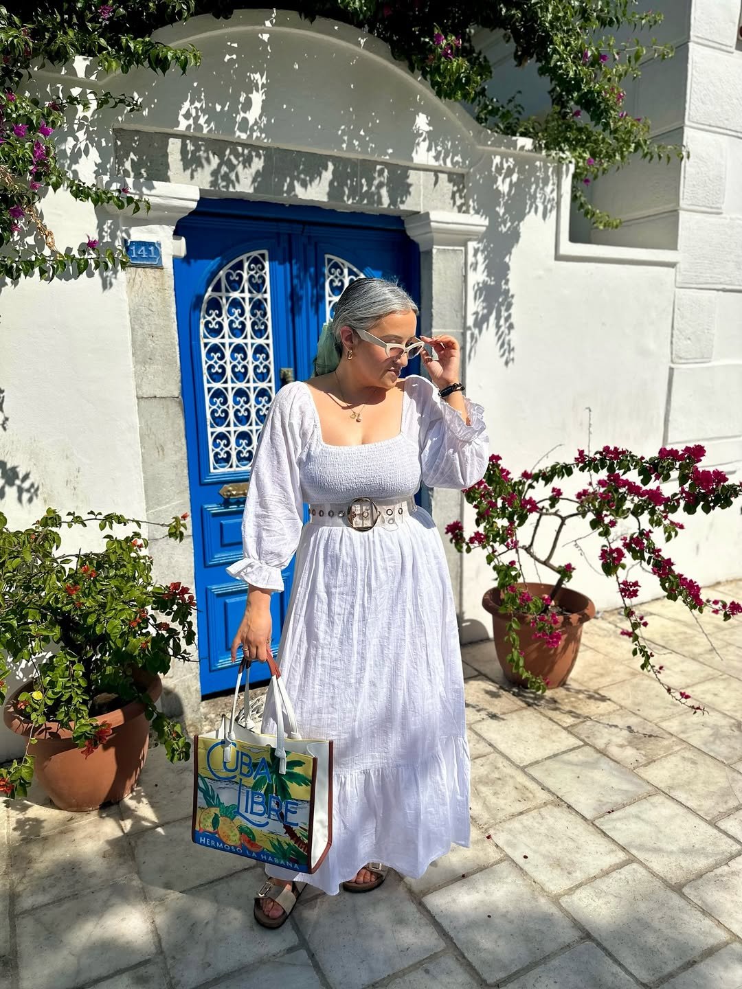 Mulher com fios brancos e vestido branco segura bolsa colorida, em frente a porta azul e plantas.