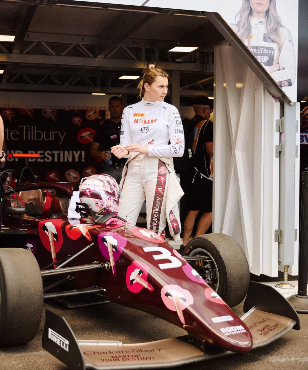Piloto ao lado de carro de corrida decorado com logotipo, em frente a uma garagem equipada, durante evento automobilístico.