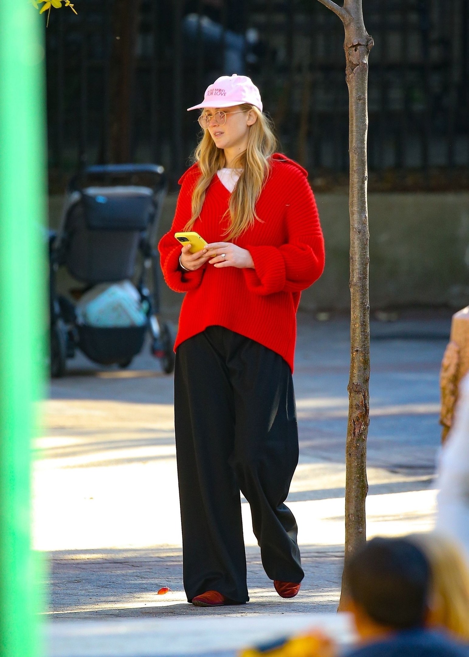 Pessoa caminhando com roupa casual, usando suéter vermelho, boné rosa e calça preta larga.
