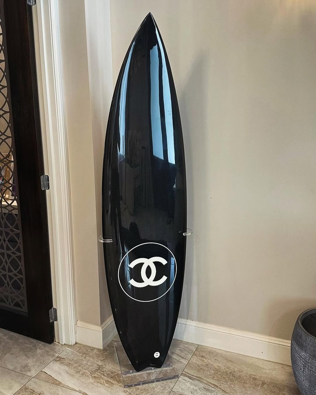 Prancha de surf preta com logotipo de moda branco, encostada na parede em um suporte transparente.