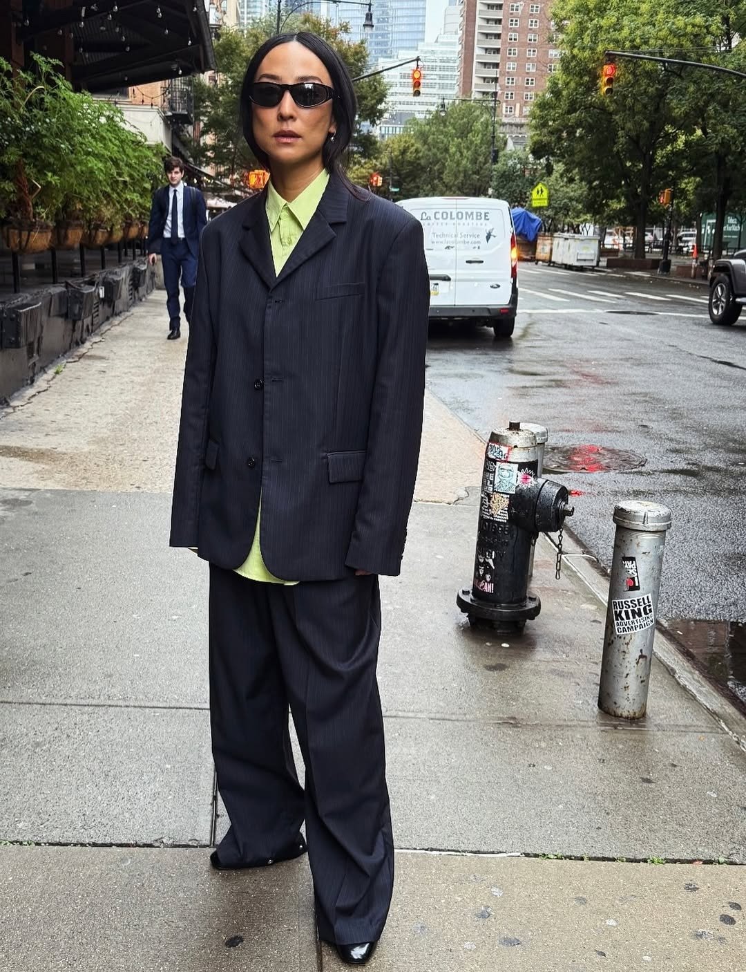O estilo da Greta Lee é uma mistura de sofisticação e ousadia. Ela usa um terno oversized em risca de giz, que traz um ar de elegância moderna. A camisa verde-limão adiciona um toque vibrante, contrastando com a paleta escura do traje. Óculos escuros mini
