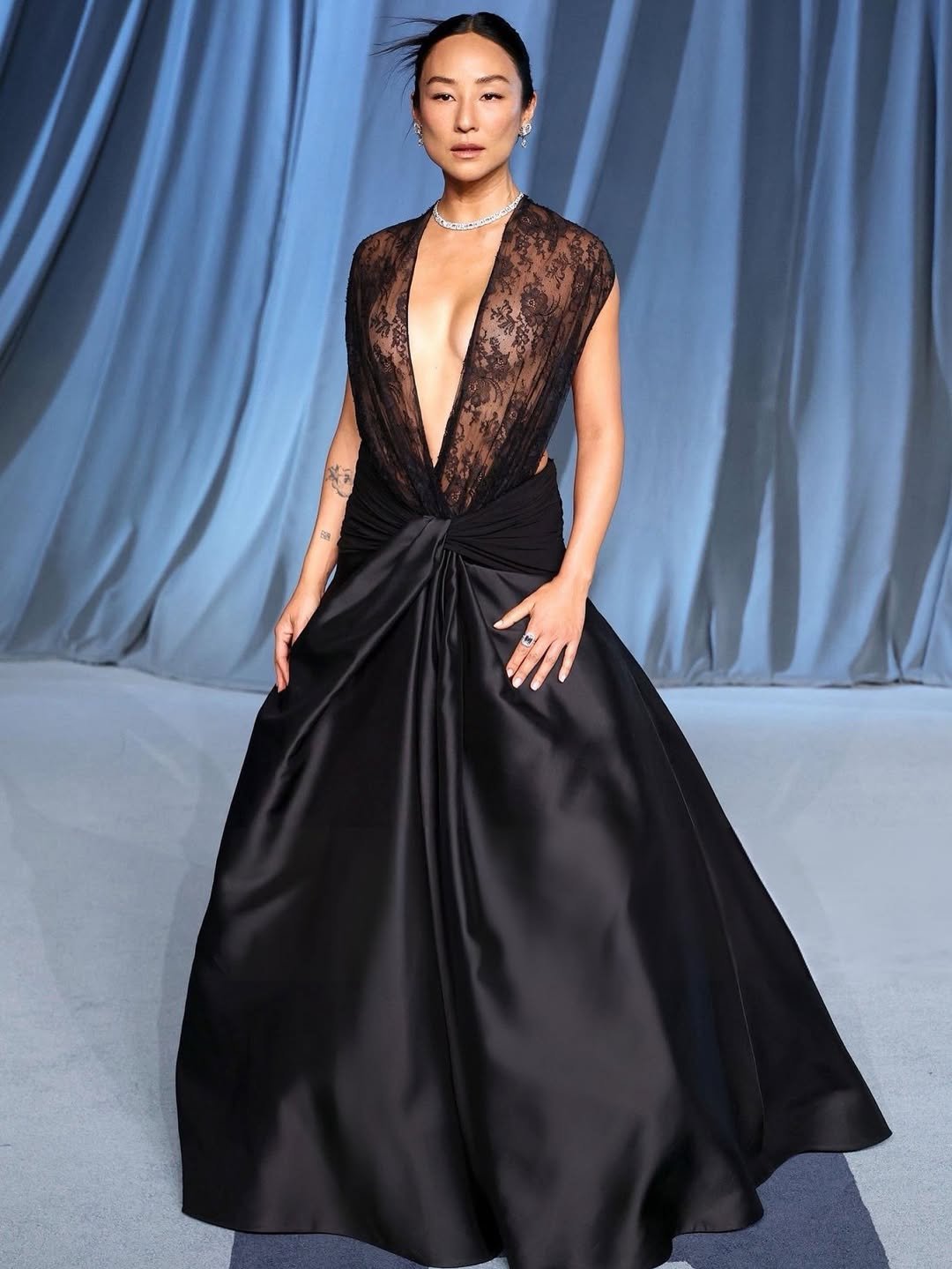 Greta Lee exibe um vestido deslumbrante em tom preto, com um decote profundo e detalhes em renda que conferem um ar sofisticado e ousado. A saia, com caimento fluido, realça a silhueta, enquanto acessórios discretos, como um colar brilhante, complementam