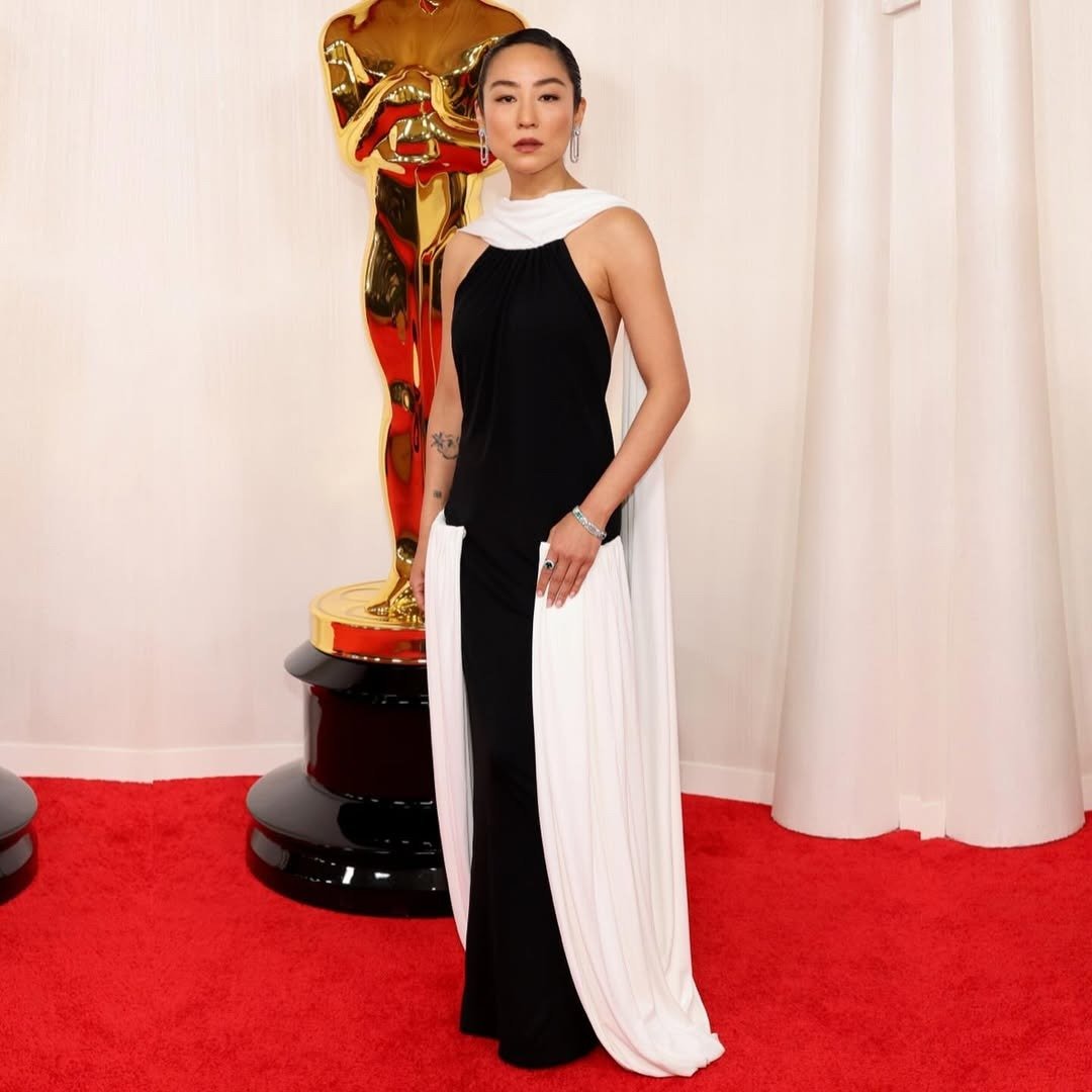 Greta Lee exibe um impressionante vestido de gala bicolor, com um design que mescla elegância e modernidade. A parte superior é preta, enquanto a parte inferior é branca, criando um contraste marcante. O vestido é complementado por uma capa que flui graci