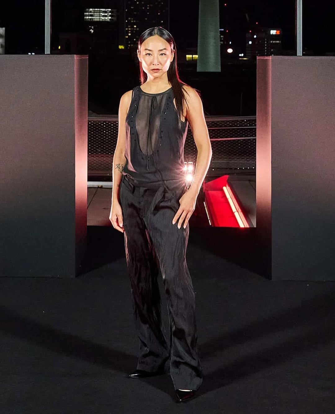 Greta Lee exibe um visual sofisticado e moderno, destacando-se em um conjunto preto de alfaiataria. A blusa, com transparência sutil e detalhes em botões, traz um toque de ousadia. As calças de corte reto complementam o look, enquanto os sapatos de salto