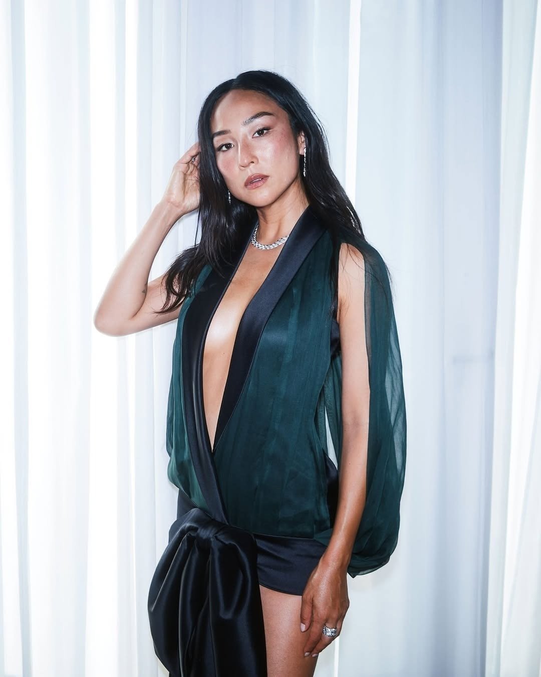 Greta Lee exibe um estilo marcante com um look ousado. Ela usa uma blusa transparente em tons de verde escuro, com decote profundo e mangas volumosas, combinada com um short preto que destaca sua silhueta. Acessórios como um colar de corrente e brincos el