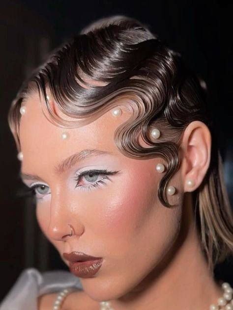 O penteados de Halloween apresentado destaca ondas glamourosas e bem definidas, com mechas estilizadas que emolduram o rosto. Pequenas pérolas são incorporadas, adicionando um toque sofisticado e misterioso.