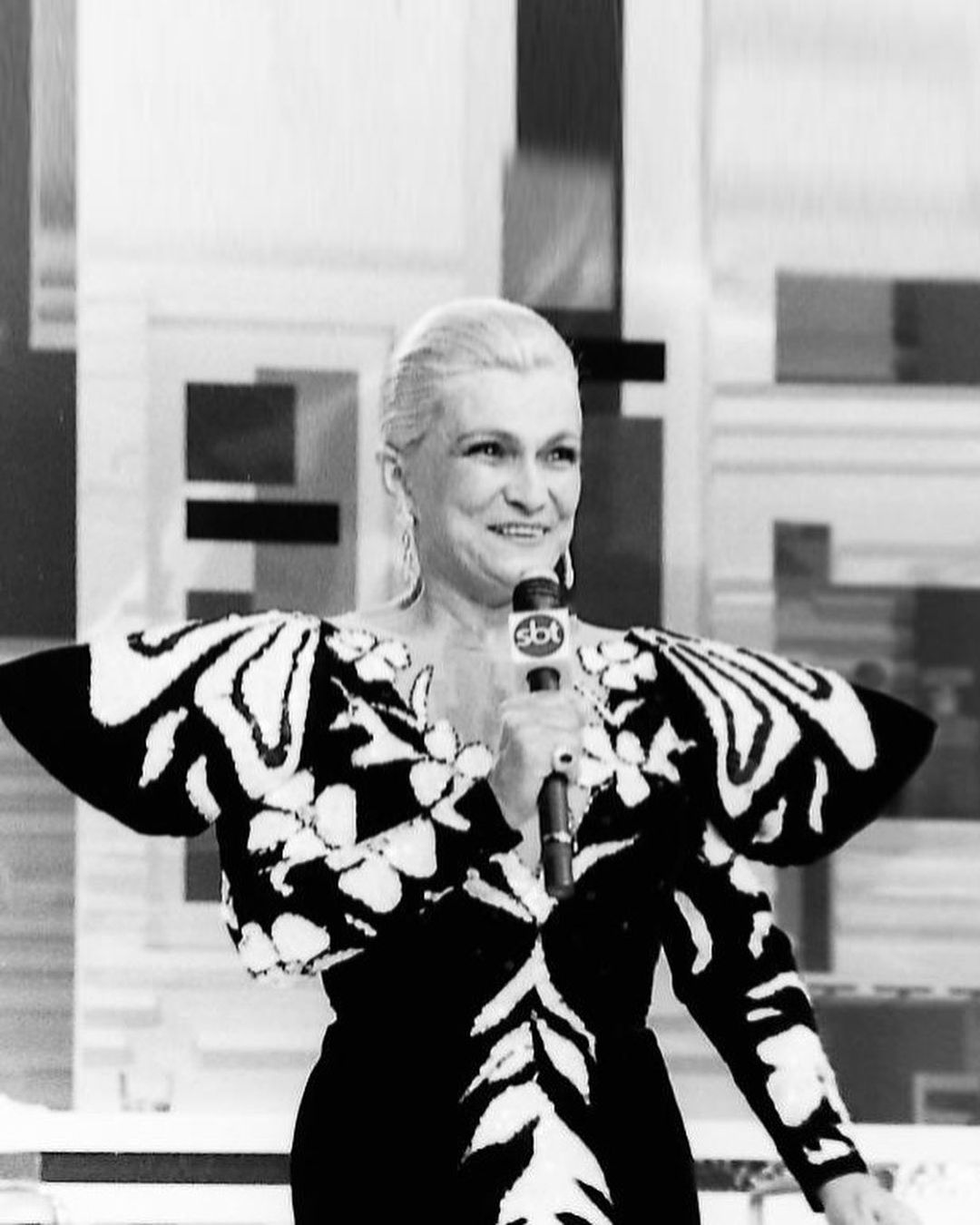 Hebe Camargo brilha em um vestido preto e branco com padrões florais e mangas amplas, criando um visual marcante e ousado. Seu cabelo platinado está preso, destacando seu rosto sorridente.
