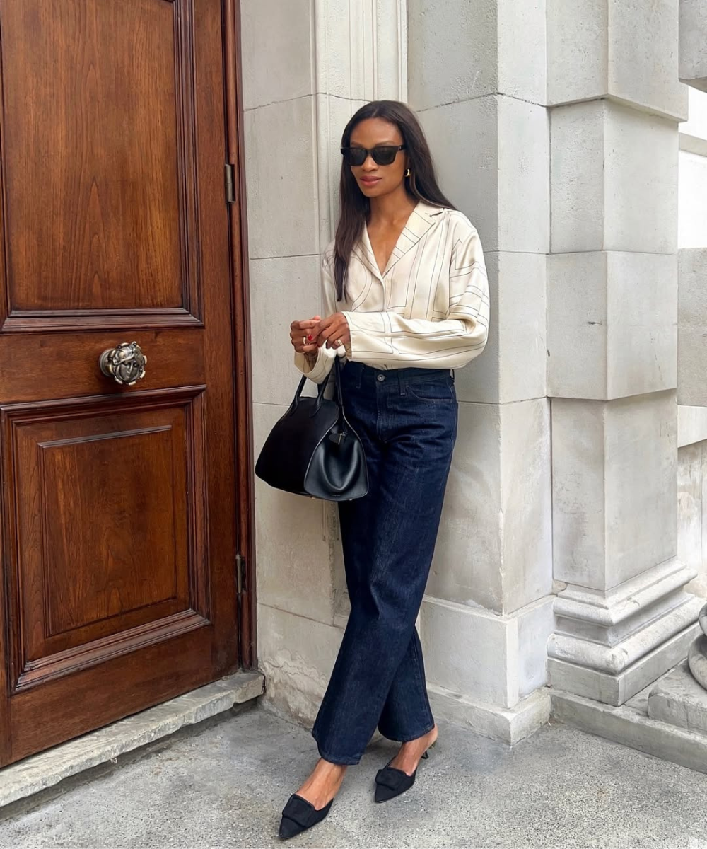 Pessoa usando camisa bege com listras, calça jeans escura, sapatos pretos e bolsa preta. Óculos escuros completam o visual.