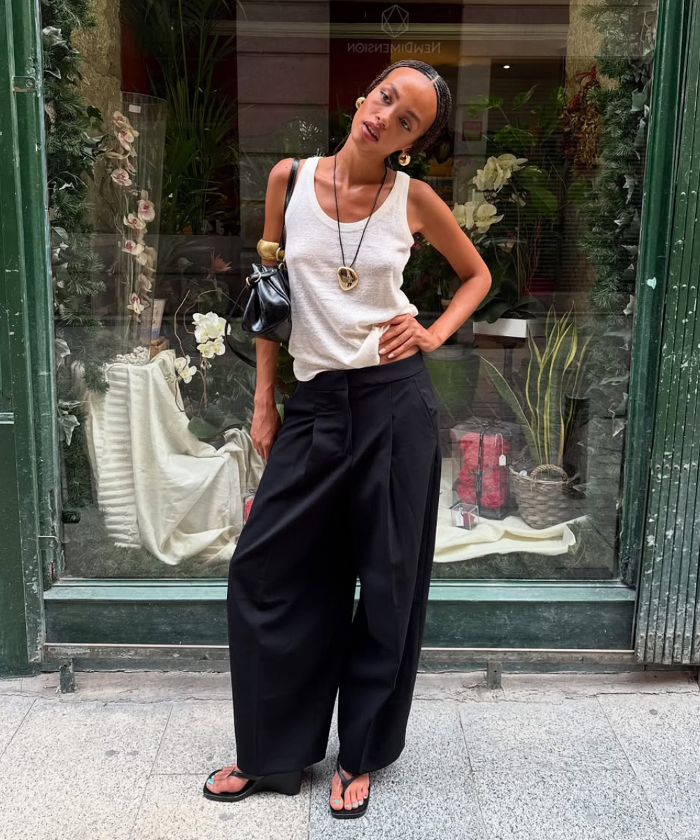 Blusa branca e calça preta larga, sandálias, bolsa e colar. Ideias de look casual em frente a uma vitrine decorada.