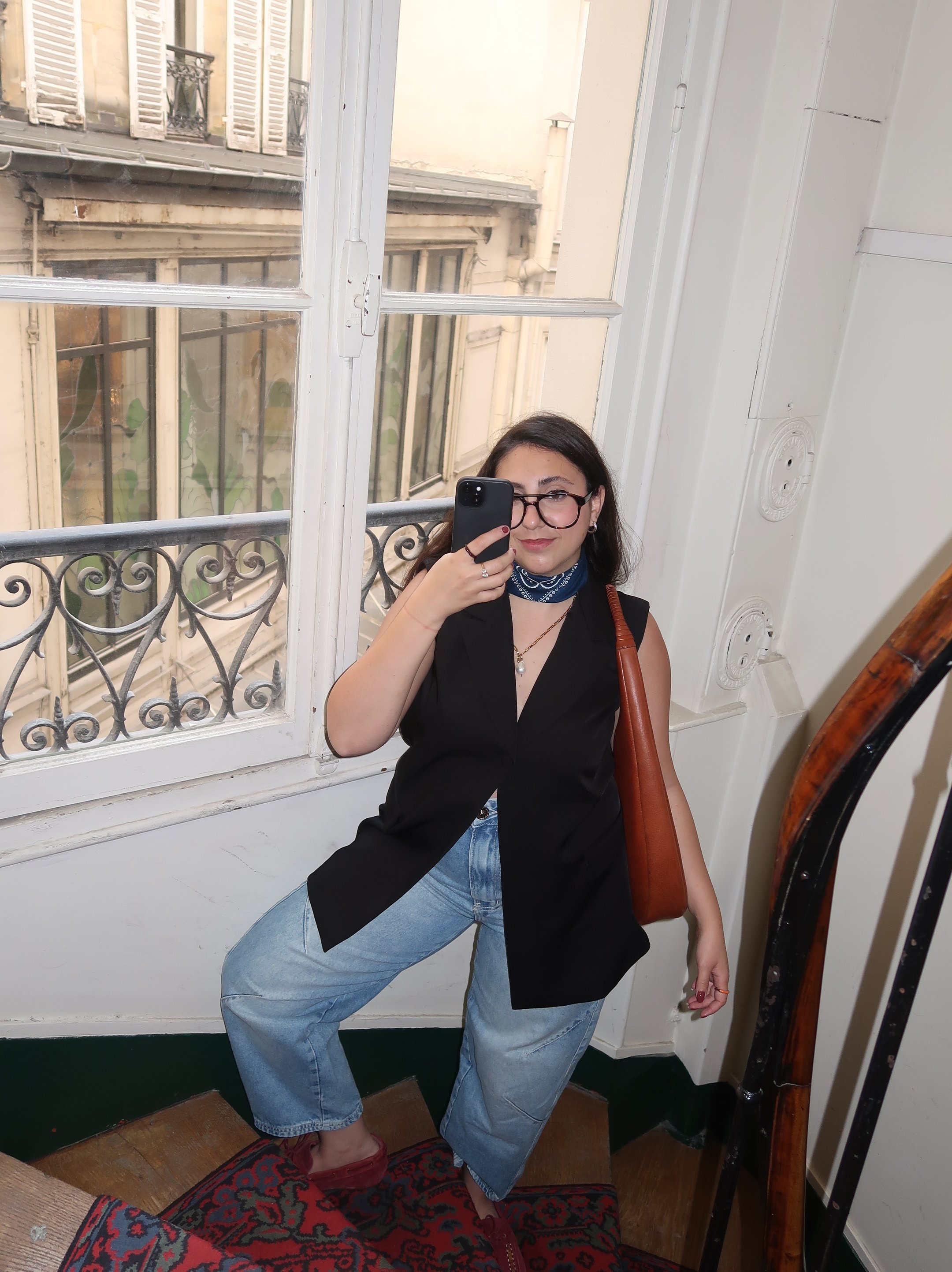 Pessoa em escada com blusa preta e calça jeans, segurando celular. Ambiente iluminado por janela vintage. Mala do Paris Fashion Week.