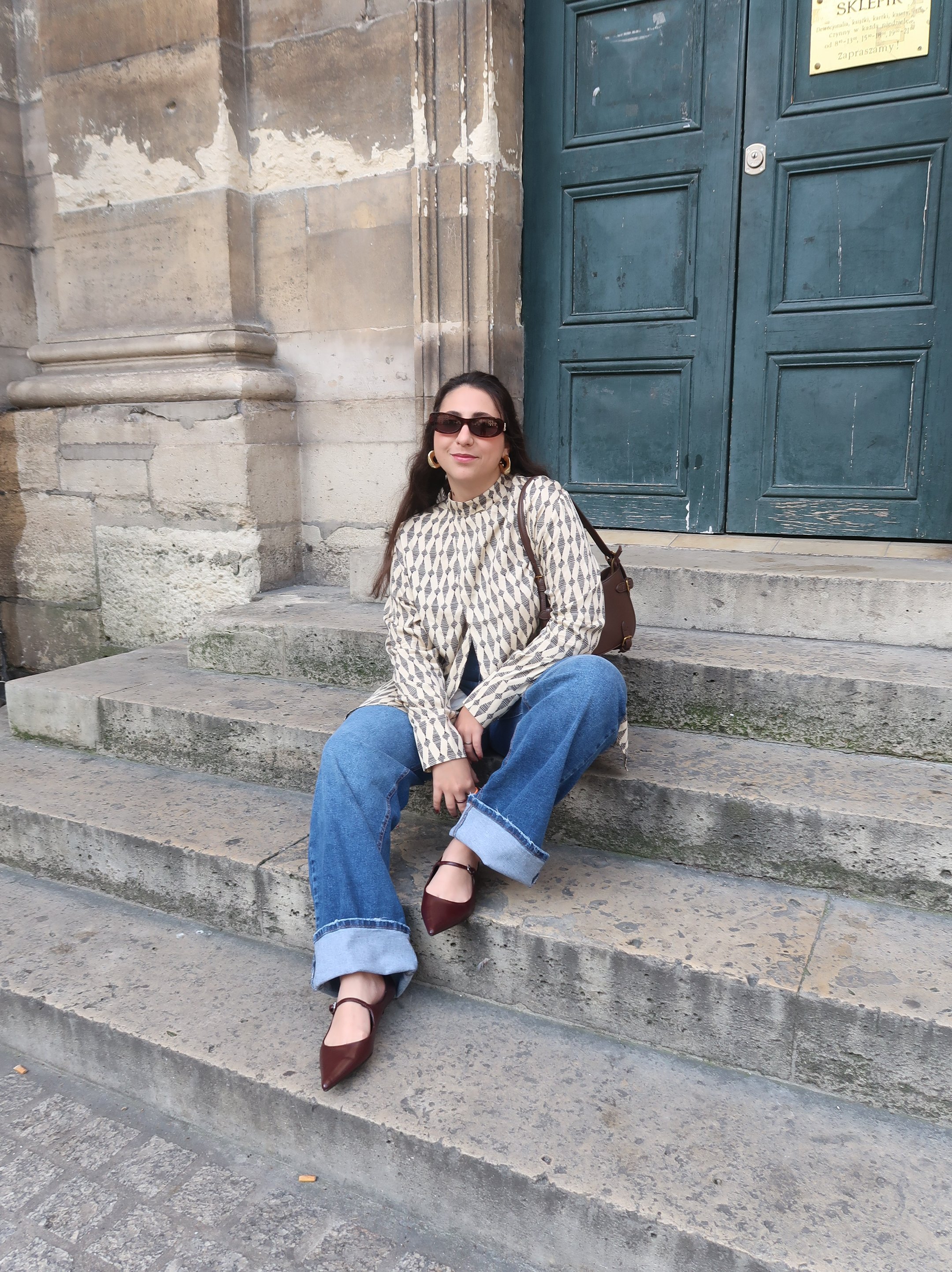Pessoa sentada em escadas de pedra, vestindo blusa estampada e calça jeans larga. mala do Paris Fashion Week