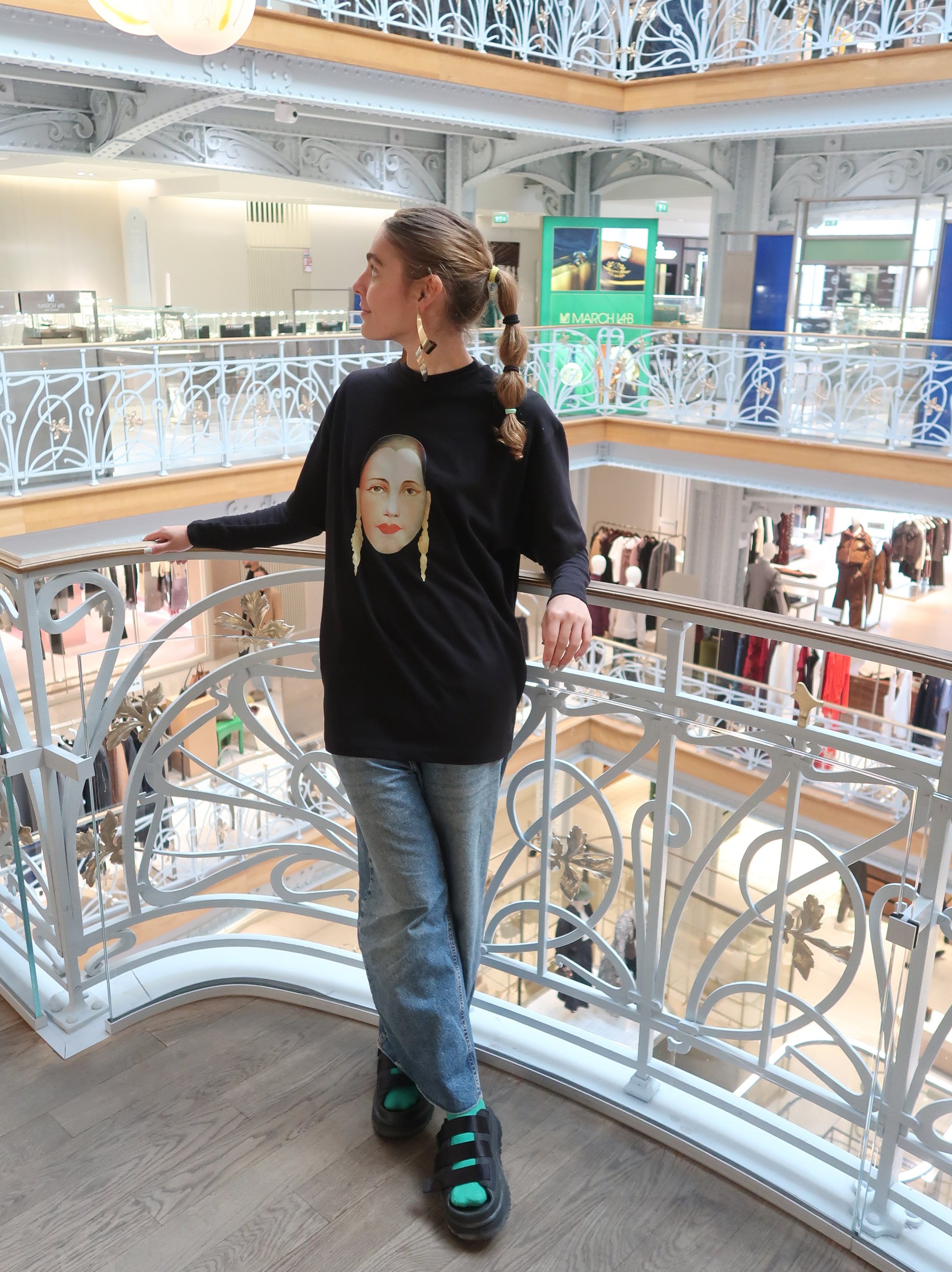 Pessoa com moletom preto e estampa, calça jeans e sandálias em shopping iluminado. Ambiente sofisticado Mala do Paris Fashion Week.