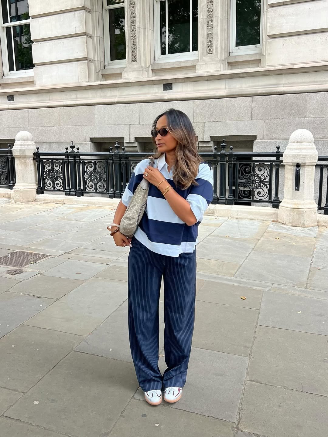 Pessoa usando blusa polo listrada azul e branca, calça azul e carregando bolsa, em frente a prédio clássico.