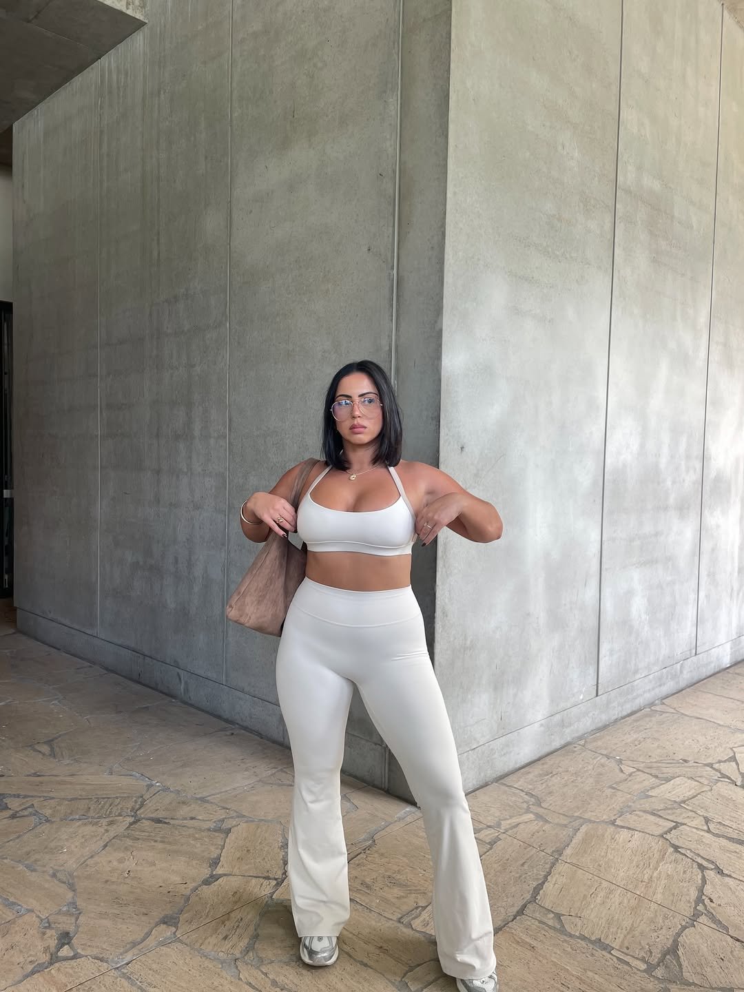 Mulher com legging flare branca e top, segura bolsa. Ao fundo, parede de concreto.