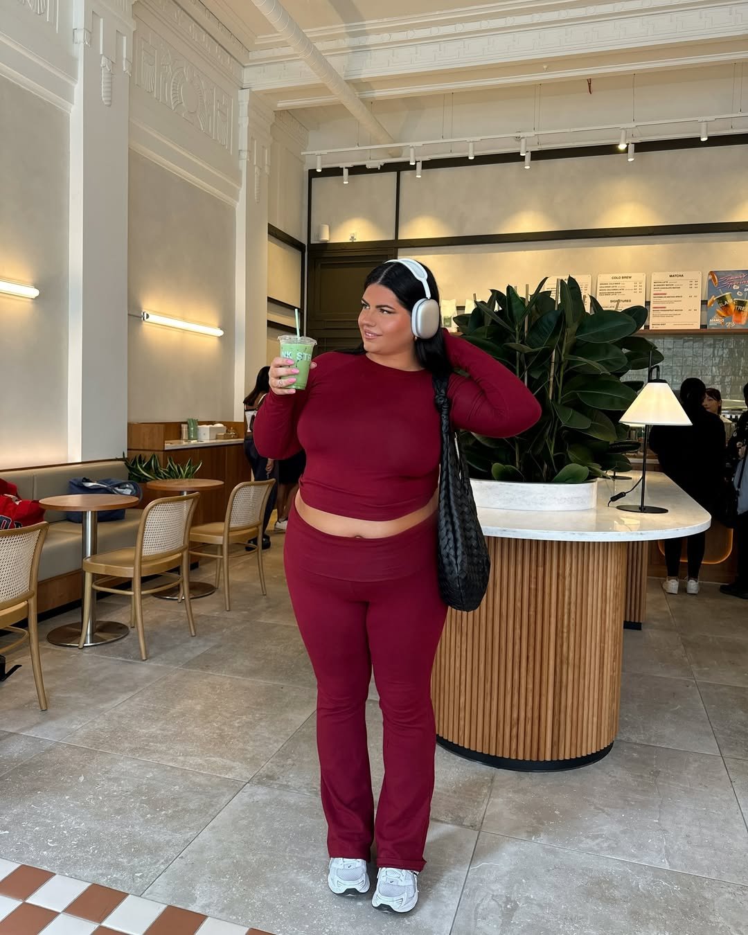 Pessoa usando roupa vinho, incluindo uma blusa e legging flare, segurando bebida verde em cafeteria moderna.
