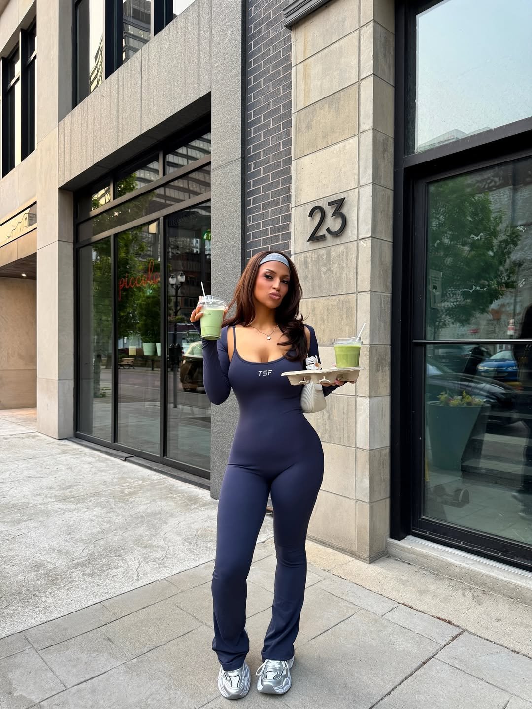 Mulher em frente a um edifício, vestindo macacão azul com legging flare, segurando bebidas verdes.