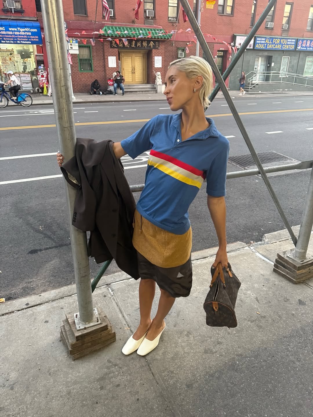 Pessoa loira em pé na calçada, vestindo uma camisa azul com listras coloridas. Look casual e moderno.