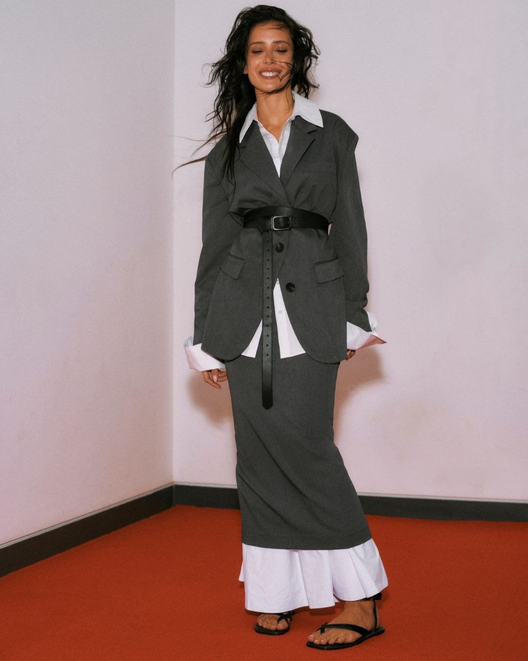 A coleção Renner + Livia traz um look sofisticado e moderno, destacando uma combinação de blazer cinza e vestido longo, que mescla elegância e conforto. O cinto acentuando a cintura adiciona estrutura à silhueta. A modelo exibe um sorriso confiante, compl