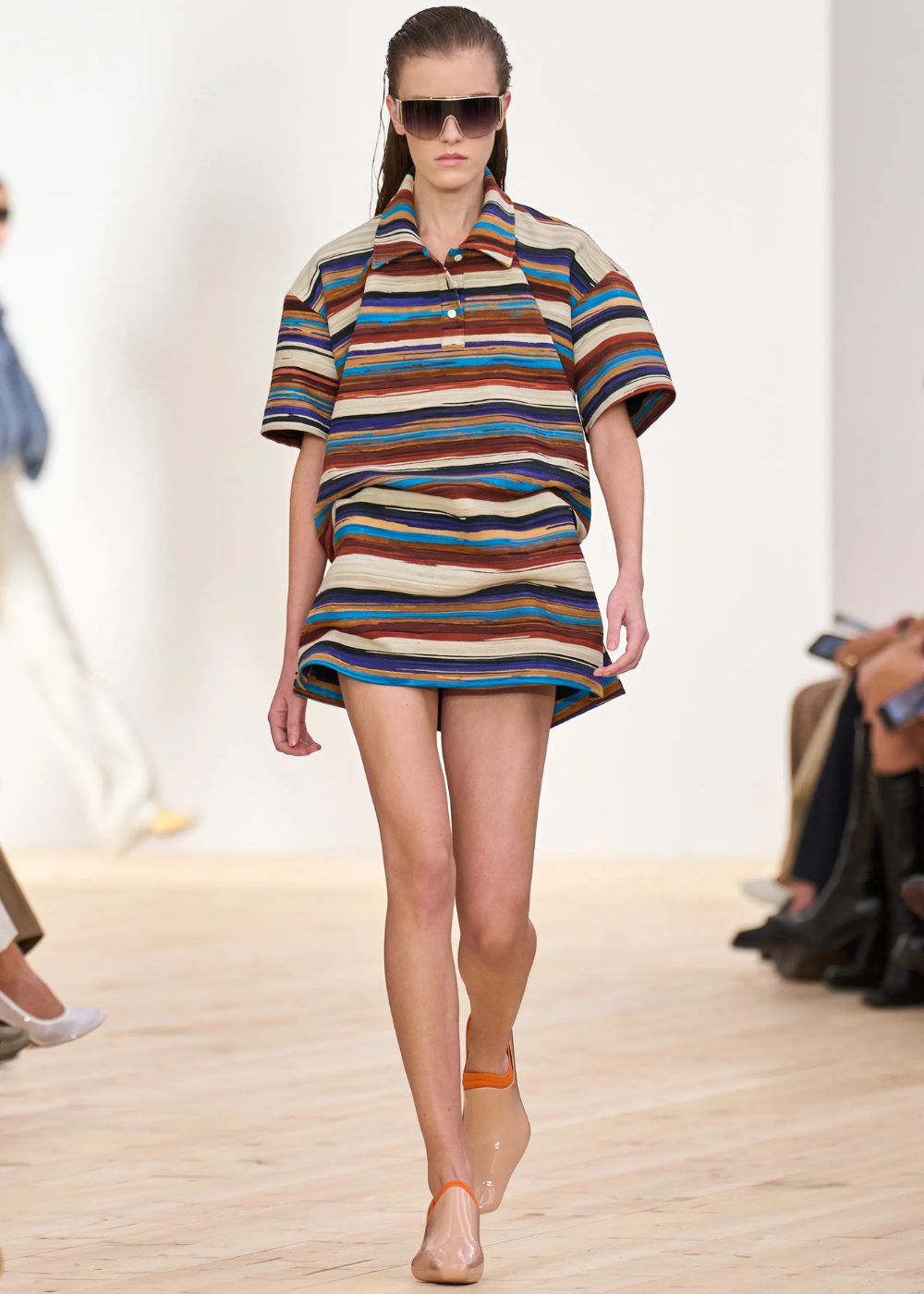 Modelo veste conjunto com blusa polo listrada, mangas curtas e colarinho, em passarela de desfile de moda.