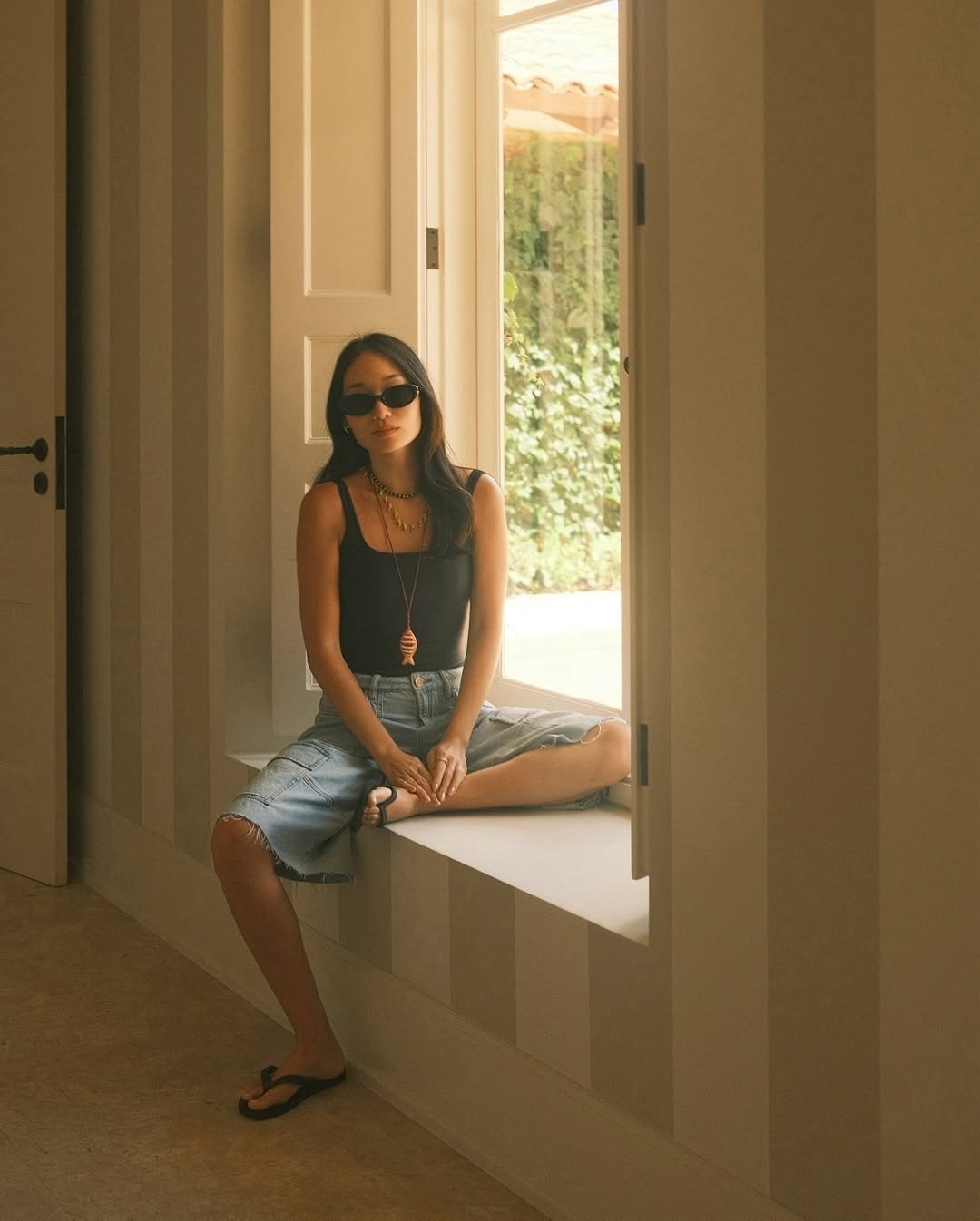 Mulher usando look para barzinho à noite com bermuda jeans, sentada em um parapeito, com um top preto justo que destaca sua silhueta.