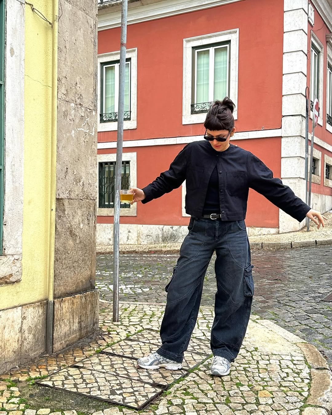 Mulher em rua de paralelepípedo usa look com calça preta cargo, jaqueta cropped e tênis cinza com branco.