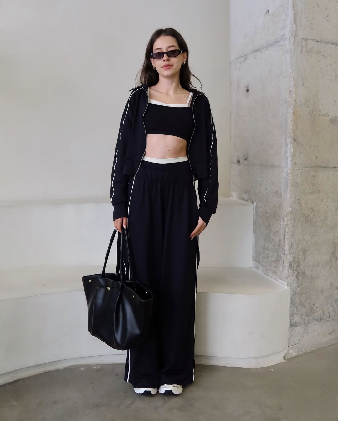 Mulher com look esportivo usa calça preta larga com listras laterais, top cropped, jaqueta combinando e bolsa preta grande.