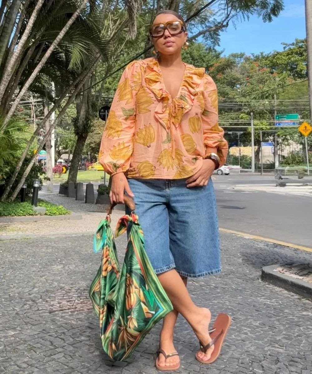 Mulher em passeio usando look com bermuda jeans e bata. A blusa tem estampa floral em tons de laranja e amarelo.