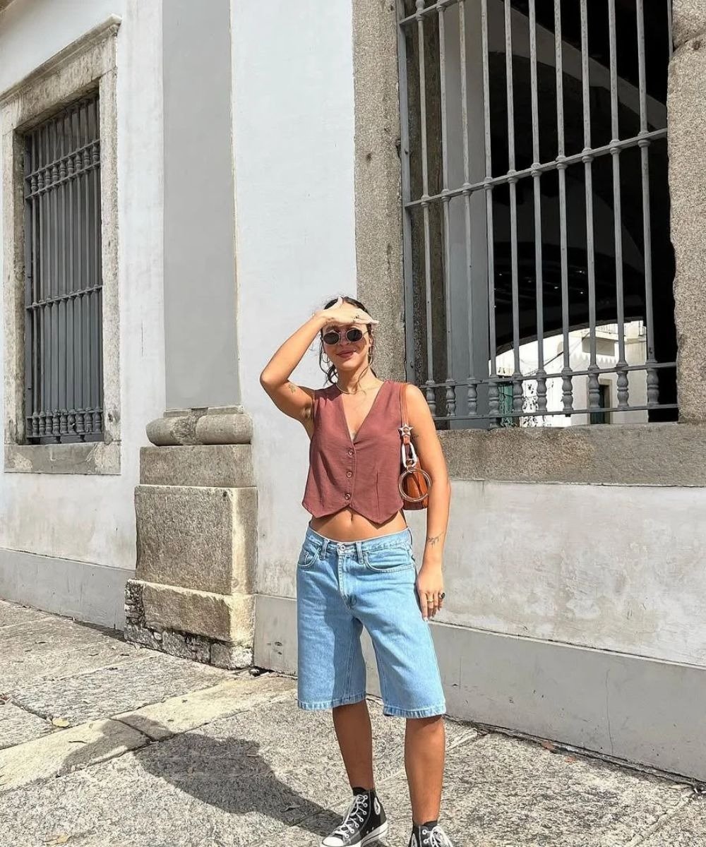 Mulher usando look com bermuda jeans feminina e tênis, complementando seu visual com um colete cropped em tom terroso.