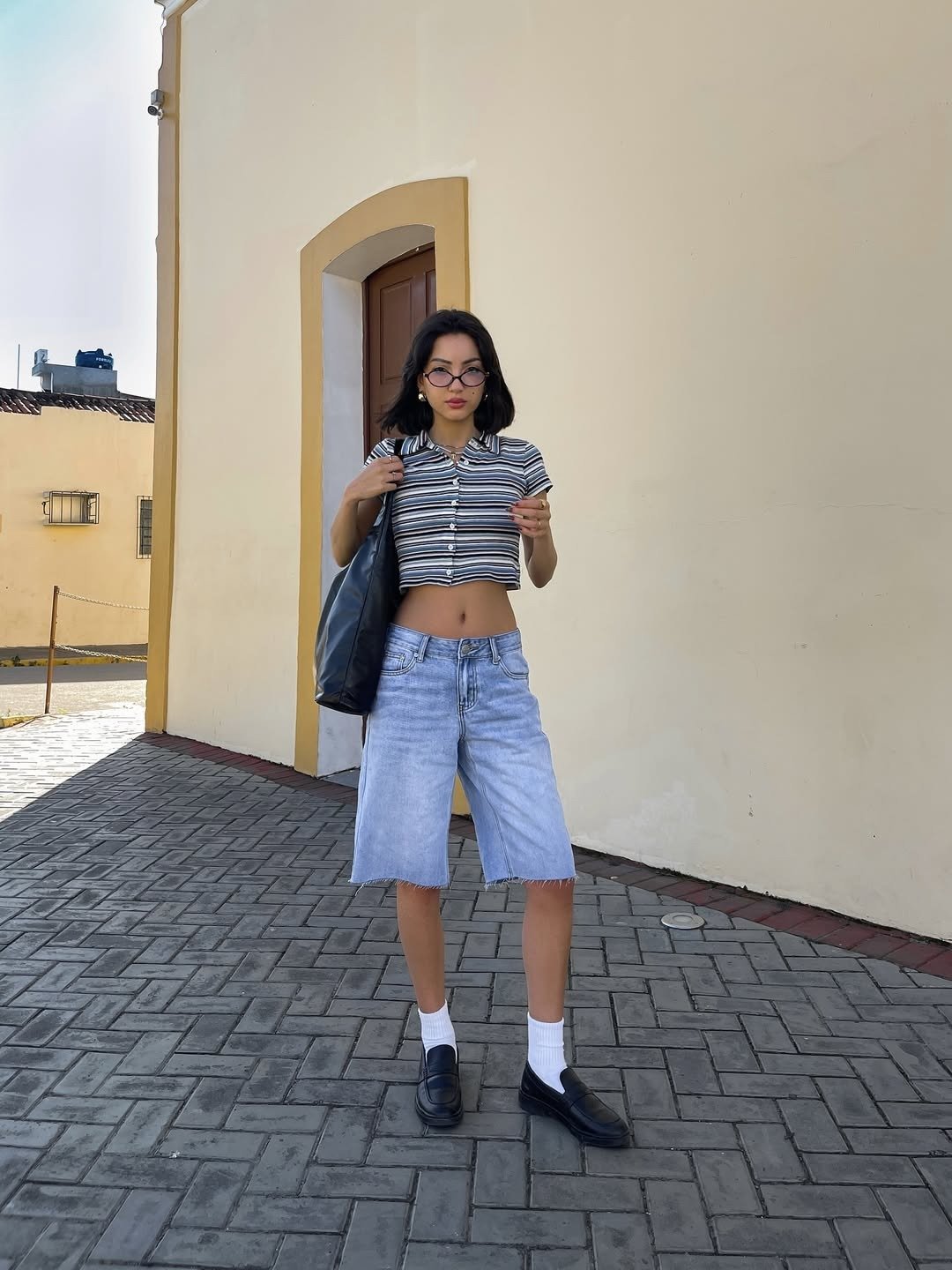 Mulher usando look com bermuda jeans para o verão com uma blusa cropped listrada em preto e branco, que destaca a cintura.