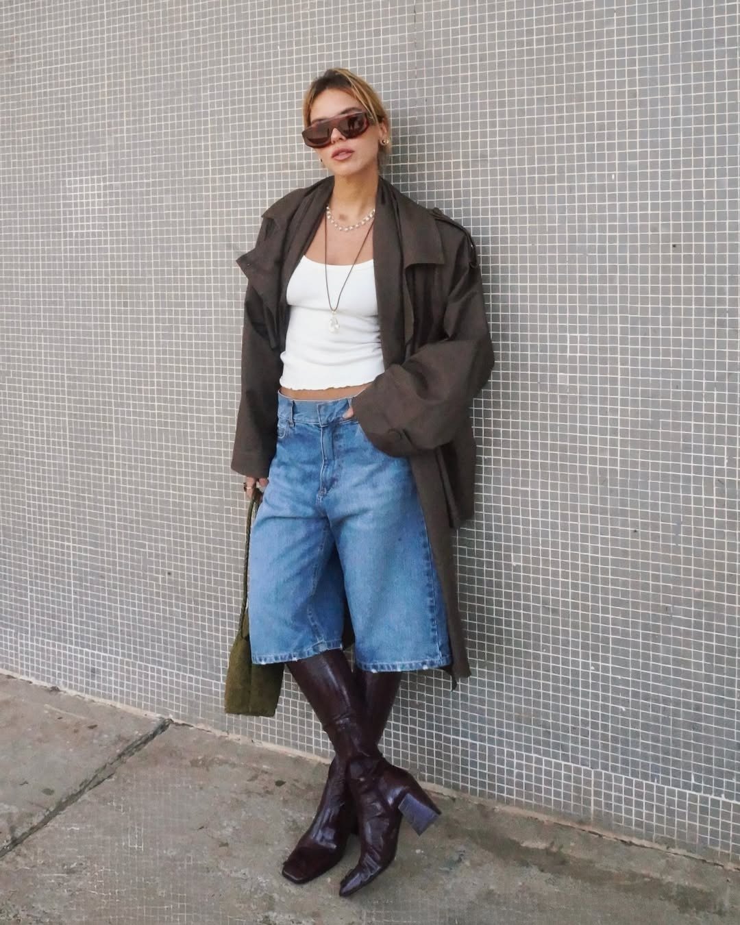 Mulher loira encostada na parede usando look com bermuda jeans para dias frios. Ela veste uma blusa branca justa, acompanhada de um trench coat.
