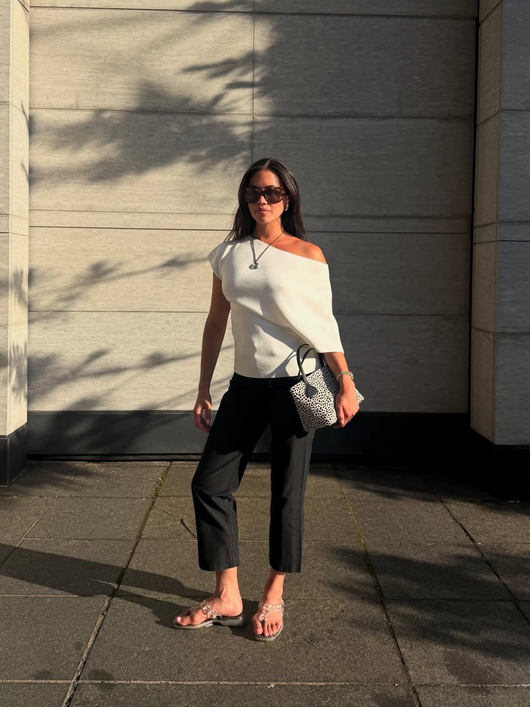 Mulher posa ao sol usando look com calça capri preta, blusa branca de um ombro só, sandálias transparentes e bolsa estampada.