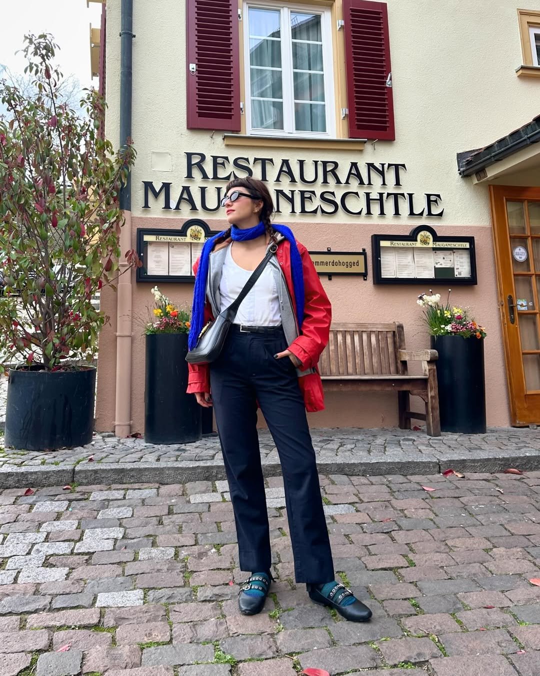 Mulher em frente a restaurante usa look com calça preta reta, camiseta branca, jaqueta vermelha e lenço azul no pescoço.