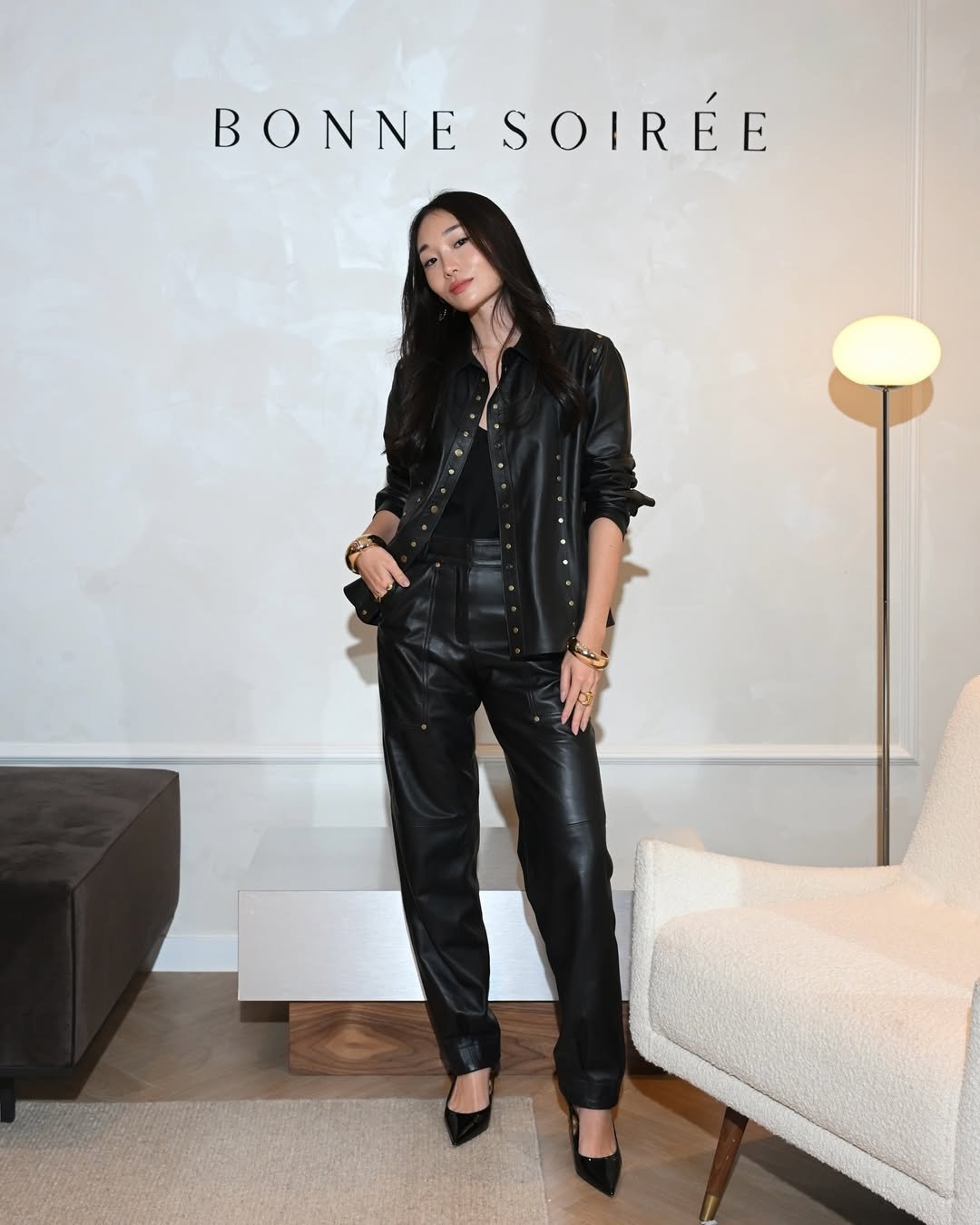 Mulher posa em evento com look sofisticado, usando calça de couro preta, camisa combinando com botões dourados e scarpin preto.