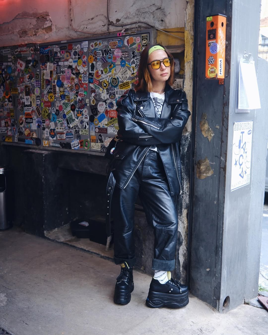 Mulher com estilo alternativo usa look com calça de couro preta, jaqueta combinando, camiseta estampada e tênis plataforma.